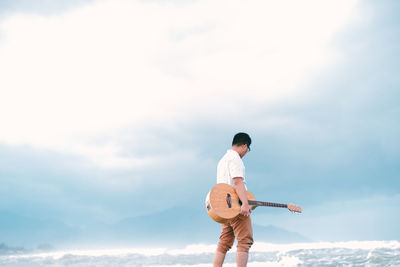 Man holding guitar walking at sea shore against sky