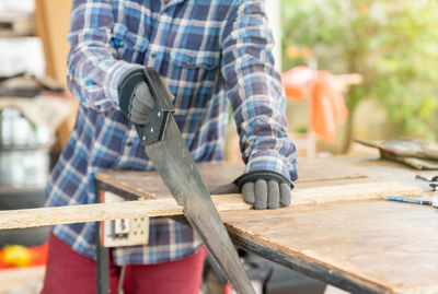 A carpenter ware gloves use hand saw to sawing wood on a table in a workshop.