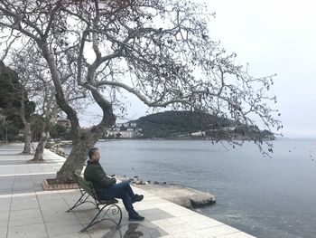 Side view of man sitting on bench by tree against sky