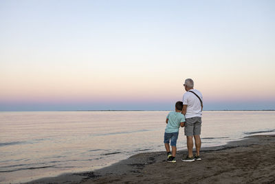 Rear view of grandfather with grandson on beach against sky during sunset