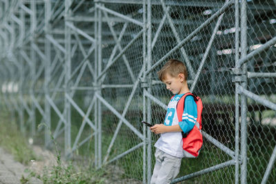Boy using mobile phone while leaning against chainlink fence