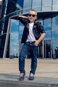 Baby boy  in sunglasses and black leather jacket smiling of the glass building in the summer,