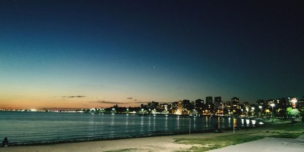 Illuminated city by sea against clear sky at night