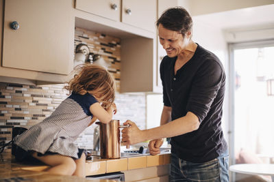 Happy father looking at daughter pushing french press while sitting on kitchen counter