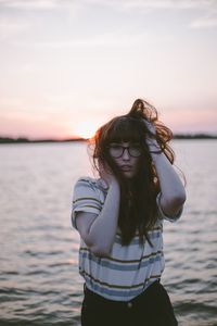 Beautiful young woman standing by sea against sky during sunset