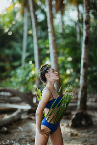 Young female in blue swimwear posing looking away with green palm leaves outfit while resting on tropical beach with lush vegetation during summer holidays in costa rica