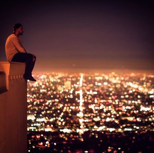 Close-up of man sitting on retaining wall against illuminated city at night