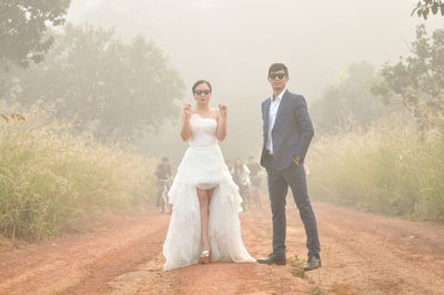 Portrait of bride and groom wearing sunglasses while standing on dirt road during foggy weather
