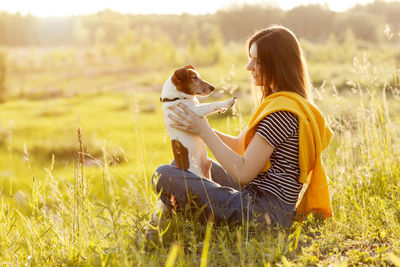 A young girl sits on the grass in the rays of the setting sun and holds her dog in her arms.