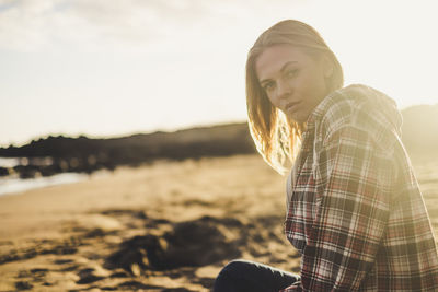 Portrait of young woman sitting at beach against sky during sunset