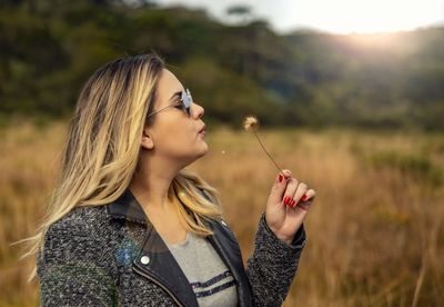 Young woman wearing sunglasses blowing dandelion on field