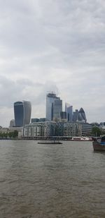 View of city buildings by river against cloudy sky