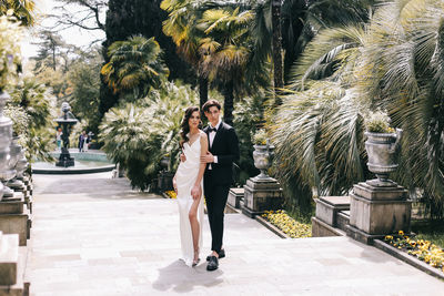 Happy lovers the bride and groom in wedding outfits walk among plants and palm trees in the old park