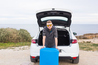 Man standing on car by sea against sky