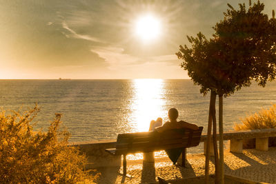 Rear view of man sitting on bench while looking at sunset over sea