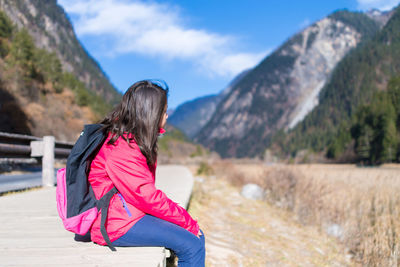 Side view of girl with backpack sitting on field against mountain