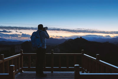 Rear view of mature man photographing with camera while standing on observation point against blue sky during sunset