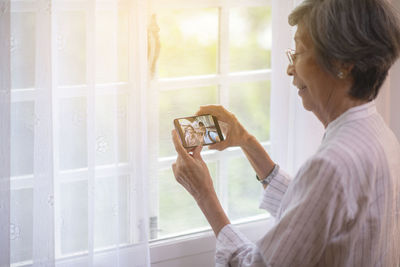 Side view of senior woman video calling while standing by window