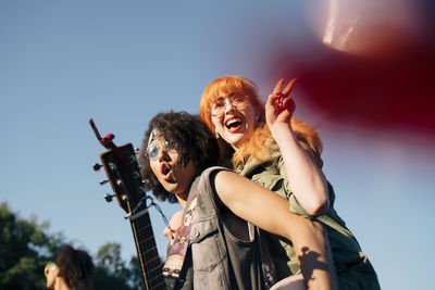 Excited man with ukulele piggybacking happy friend in event against sky