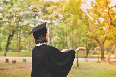 Side view of young woman in graduation gown gesturing while standing at park