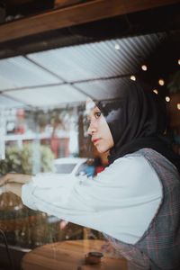 Thoughtful teenage girl wearing hijab in cafe seen through window