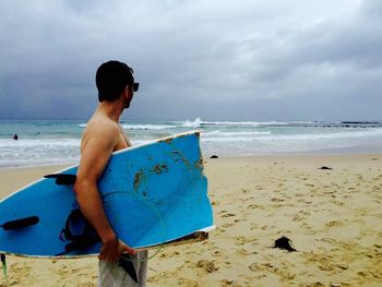 Side view of man holding broken surfboard while standing at beach against cloudy sky