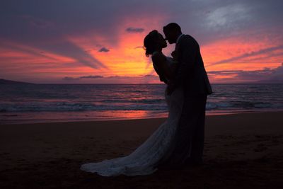 Side view of wedding couple embracing while standing on shore at beach during sunset