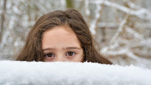 Small girl showing her eyes behind a snow layer,in the background branches covered with snow.