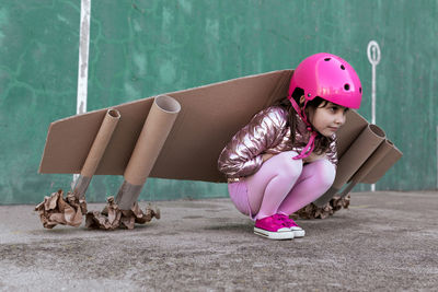 Side view of adorable little girl in cardboard plane wings and protective helmet sitting on street and looking away