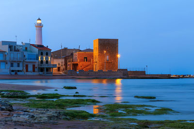 Buildings by river against clear blue sky at dusk