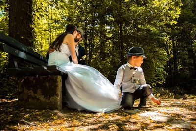 Full length of boy holding dress of bride kissing groom while sitting on park bench