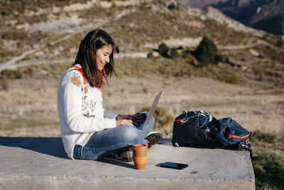 Young woman using laptop while sitting by backpack at mountain