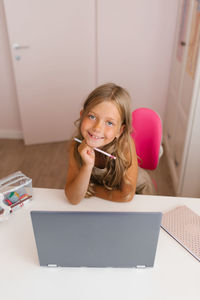 Happy schoolgirl uses a laptop, looks at the camera and studies online at home, sitting at the table