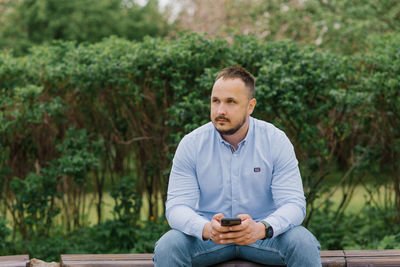 Handsome man in a blue shirt holds a smartphone in his hands outdoors, sitting on a bench making