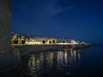 Illuminated buildings by sea against sky at night