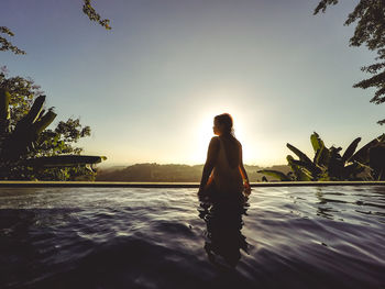 Rear view of standing by swimming pool against sky during sunset