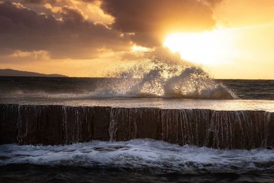 Sea waves splashing on rocks against sky during sunset