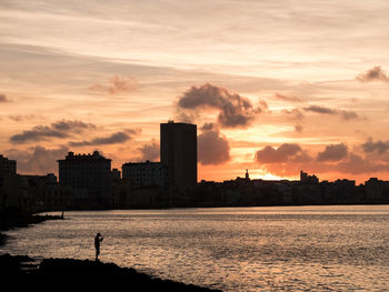 Silhouette man fishing on shore against buildings during sunset