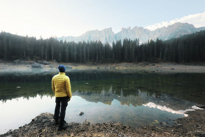 Rear view of man looking at karersee lake while standing against sky in forest during winter
