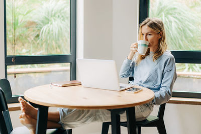 Young relaxed female freelancer drinking coffee and working remotely on laptop from home.