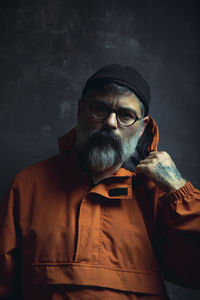 Pensive stylish tattooed adult ethnic male with gray beard in trendy hooded jacket and eyeglasses looking away against black background