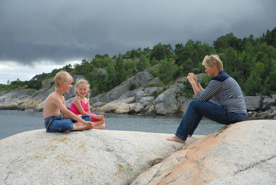 Side view of woman photographing smiling children sitting on cliff against cloudy sky