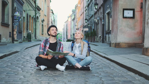 Young couple sitting on footpath amidst buildings in city