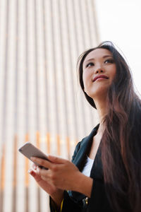 From below of happy asian female entrepreneur in smart casual style standing in street of downtown browsing on mobile phone while looking away