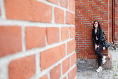 Young beautiful brunette girl in black jacket and jeans stands near wall of gothic church on summer