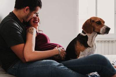 Pregnant woman smiling with male partner while dog sitting on bed