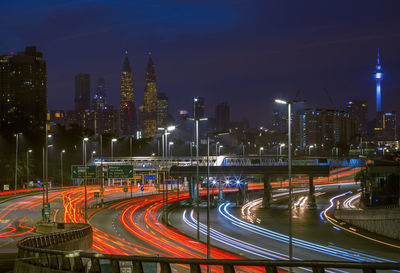 Light trails on road by buildings against sky at night