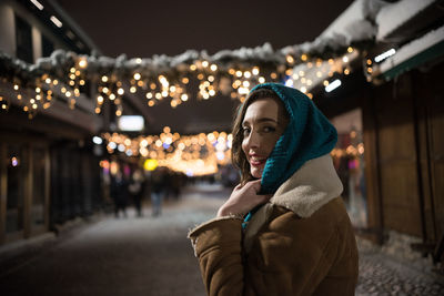 Portrait of smiling woman standing against illuminated lights during winter at night