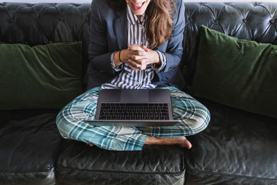 Cropped unrecognizable female entrepreneur in formal blazer and pajama pants sitting on sofa and communicating with partner via video call on laptop while working remotely from home