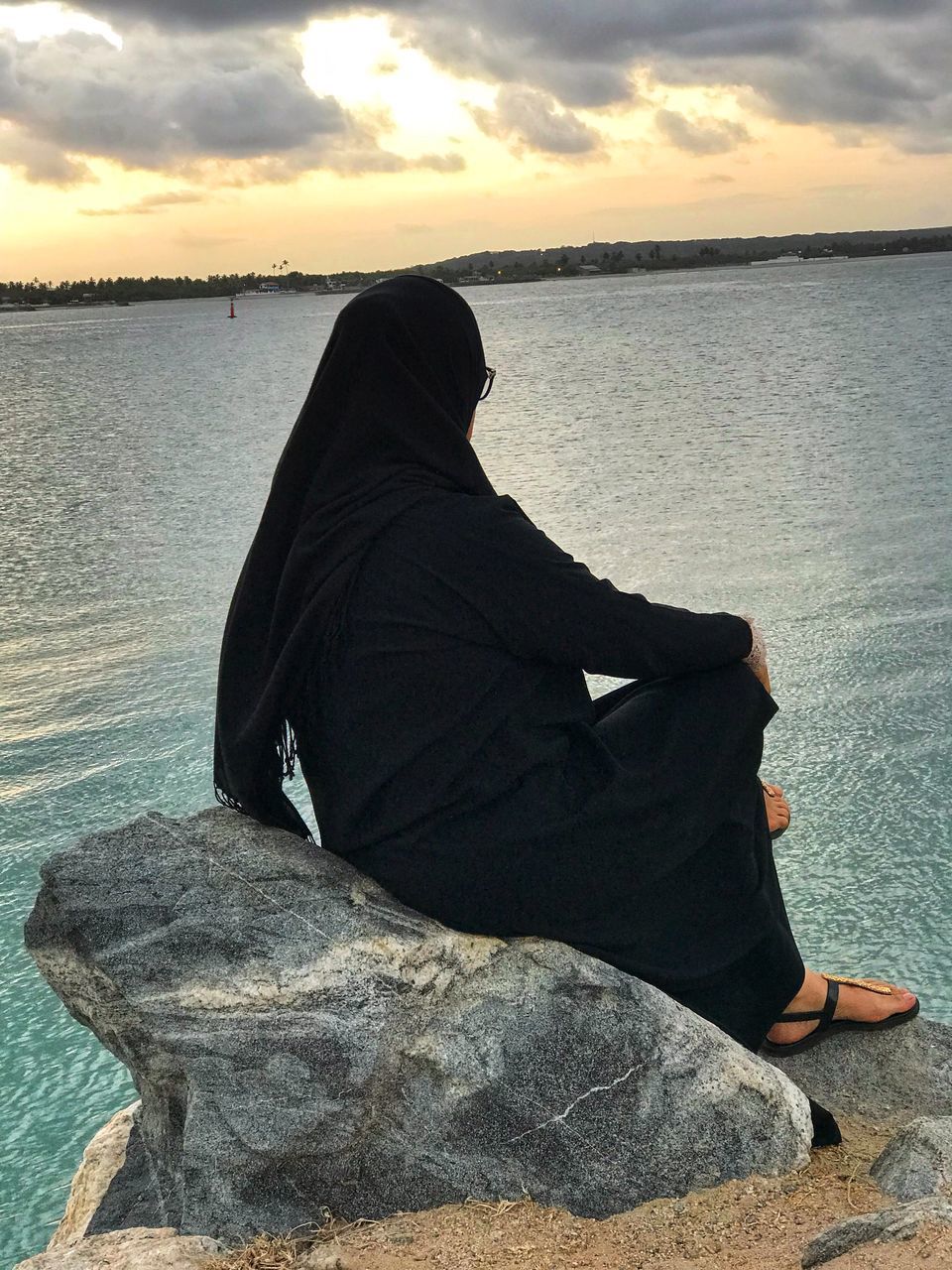 REAR VIEW OF WOMAN SITTING ON ROCK AT SHORE AGAINST SKY